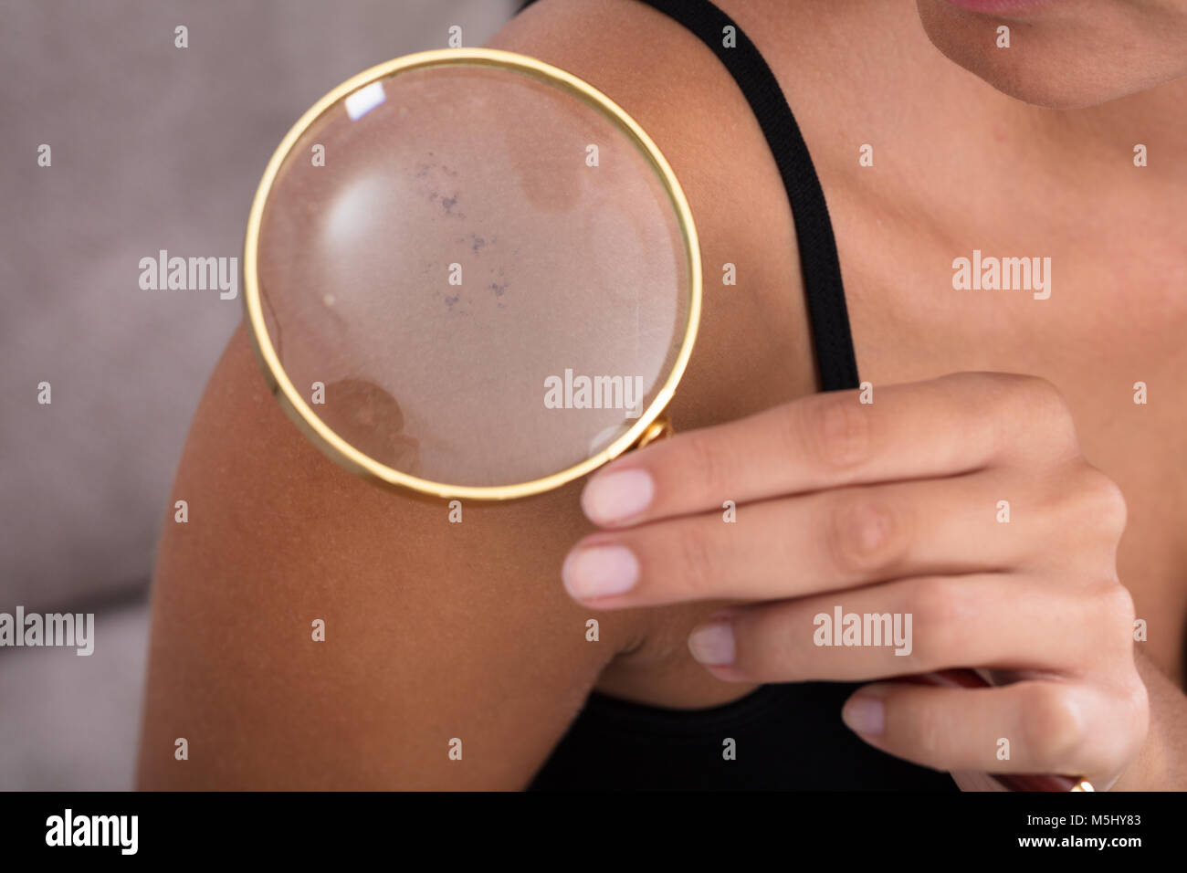 Close-up Of Young Woman Showing Skin On Shoulder Using Magnifying Glass At Home Stock Photo