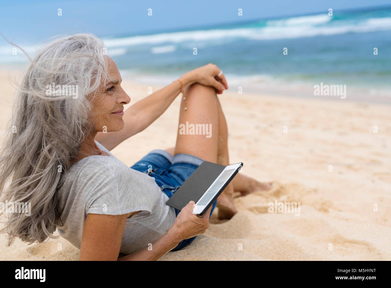 Beautiful smiling senior woman lying at the beach, holding e-book Stock Photo