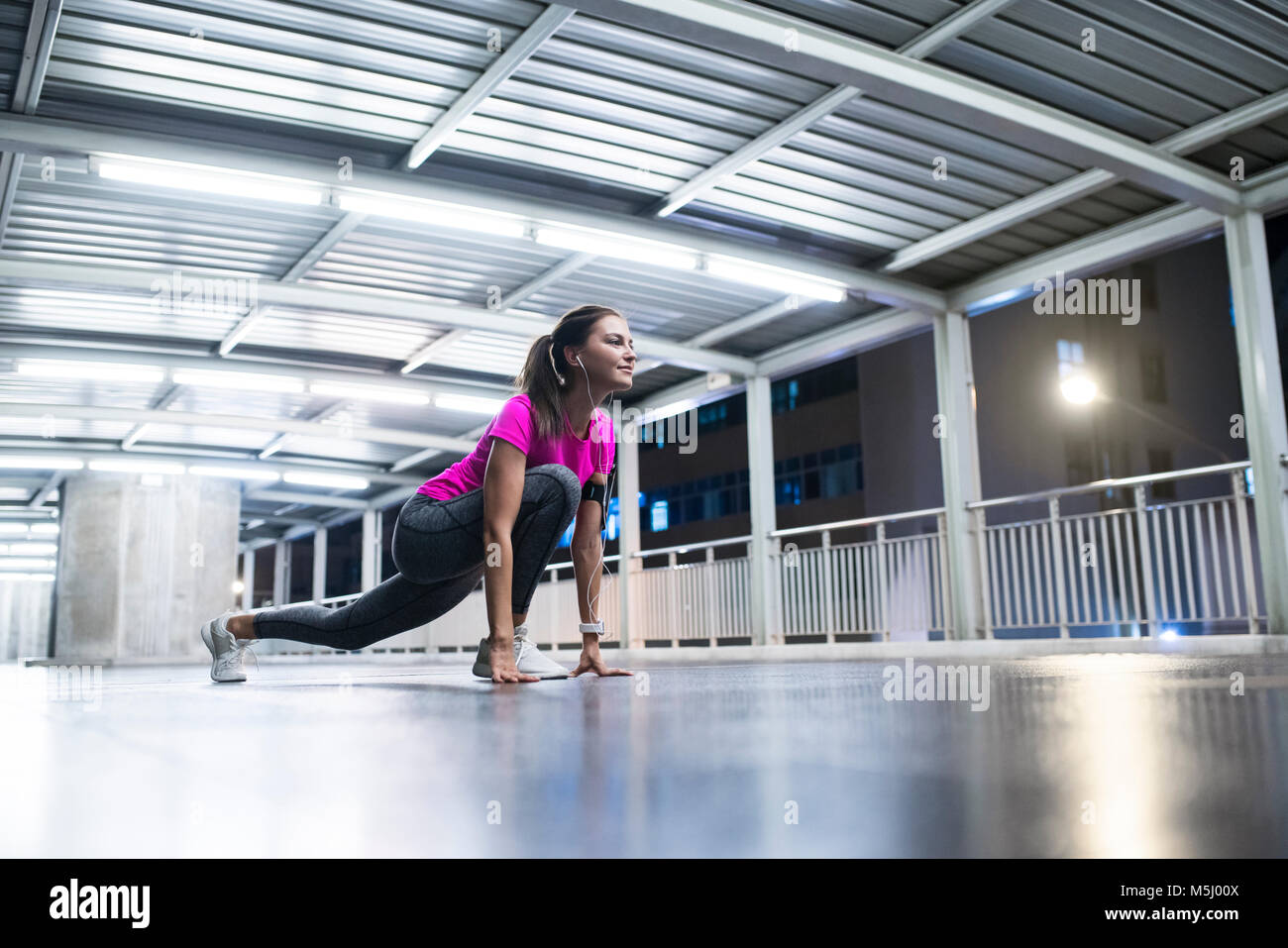 Young woman in pink sportshirt stretching in modern metro station at night Stock Photo