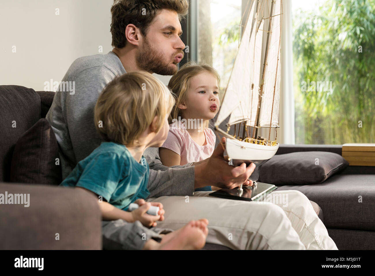 Father and children blowing into sails of toy model ship on couch at home Stock Photo