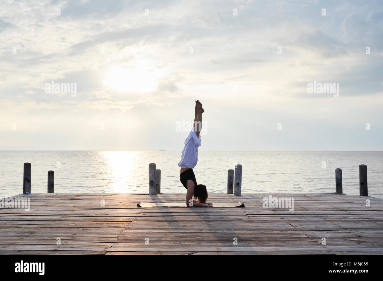 Yoga instructor in Pincha mayurasana position (elbows handstand) in a wooden bridge against sunset and sea. Kep, Cambodia. Stock Photo