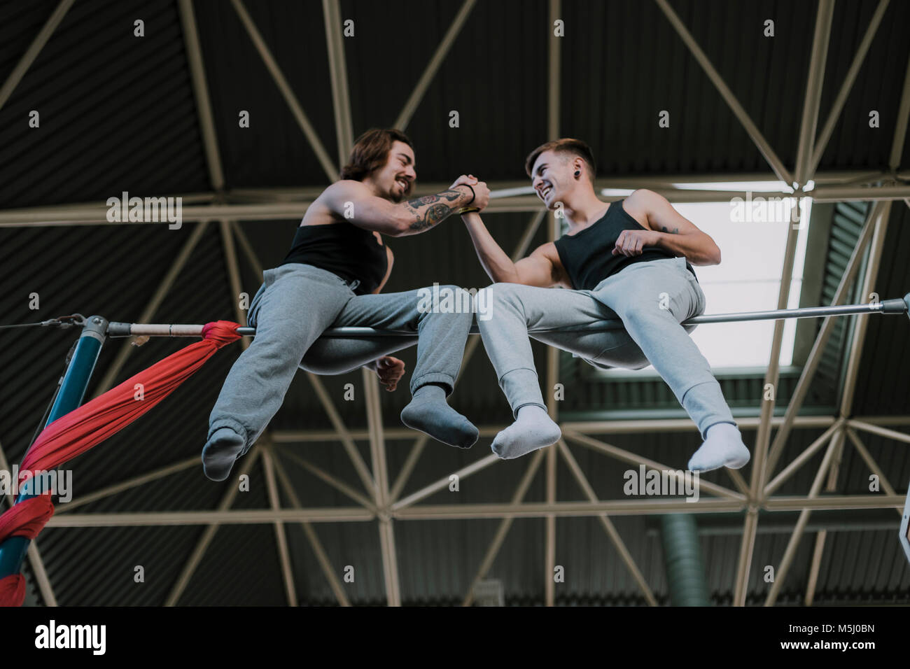 Two smiling gymnasts sitting on high bar shaking hands in gym Stock Photo