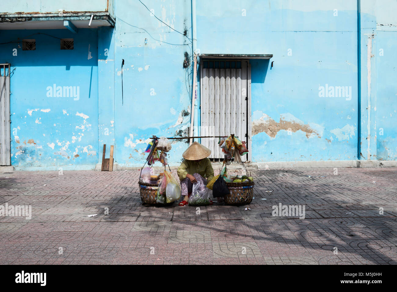 Vietnam, Ho Chi Minh, typical street vendor with vietnamese hat sitting down selling food Stock Photo