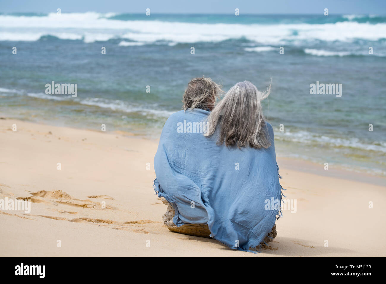 Senior couple sitting on the beach, wrapped in a blanket Stock Photo