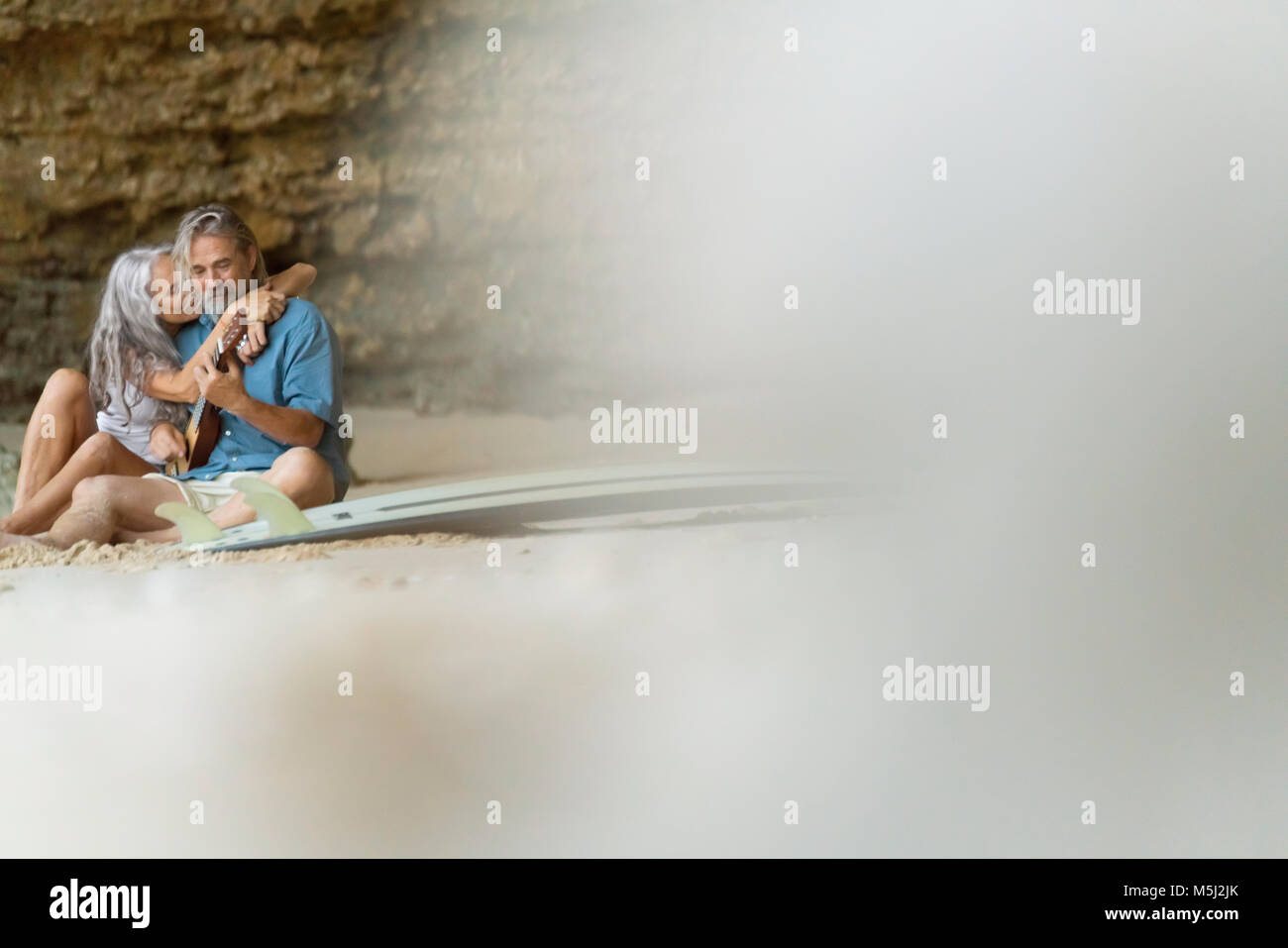 Handsome senior couple sitting on the beach, man playing guitar Stock Photo