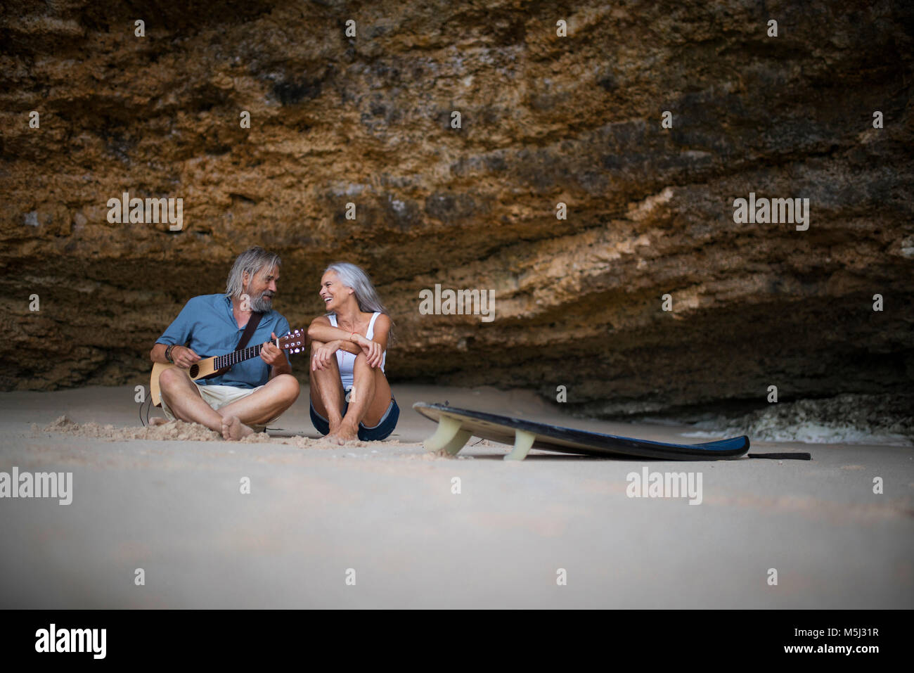 Handsome senior couple sitting on the beach, man playing guitar Stock Photo