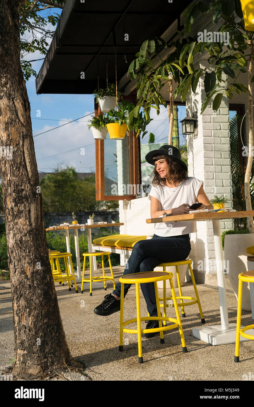 Woman with hat and smartphone sitting outside of a cafe Stock Photo