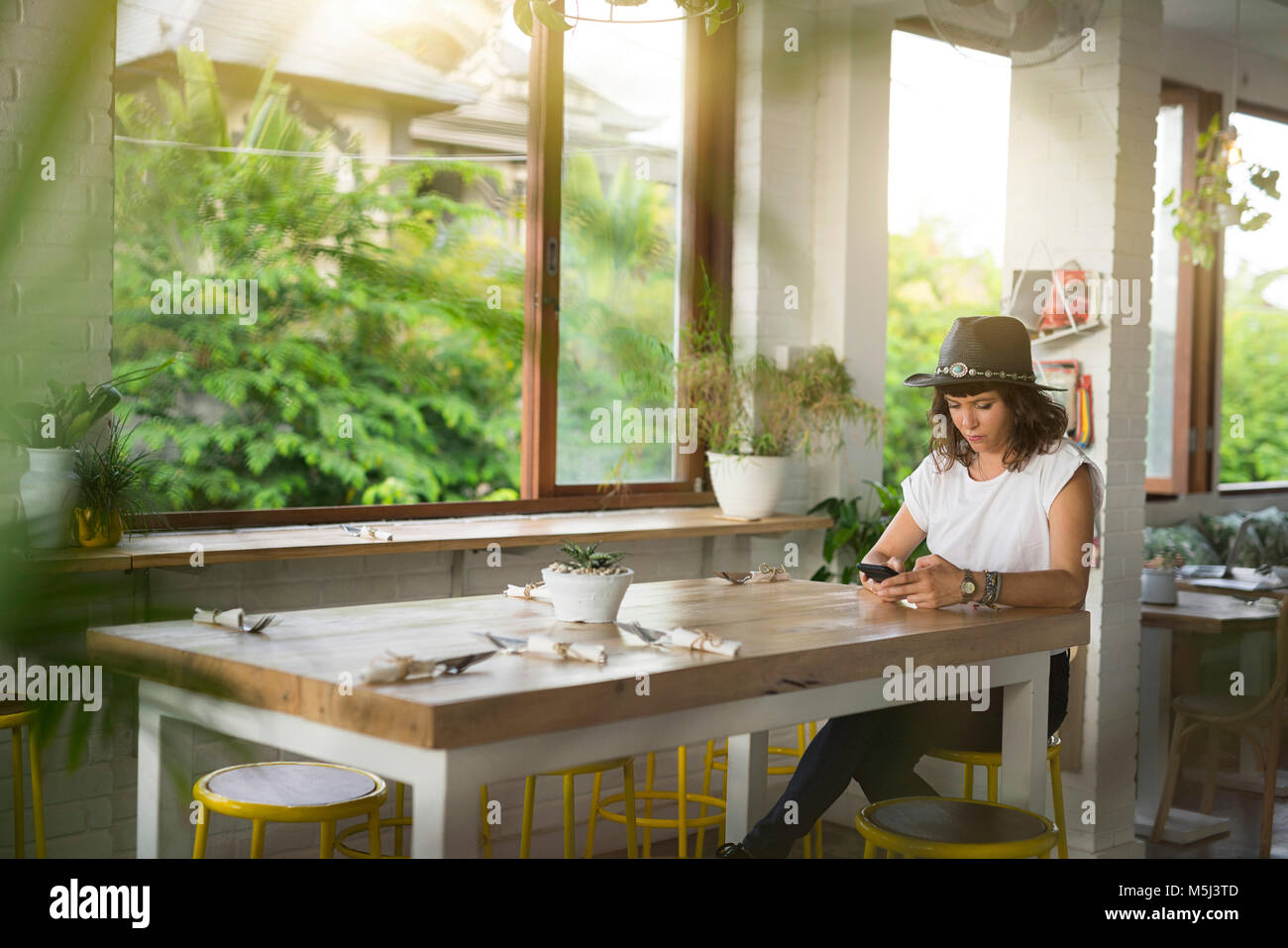 Woman with hat sitting at table in a cafe checking her smartphone Stock Photo