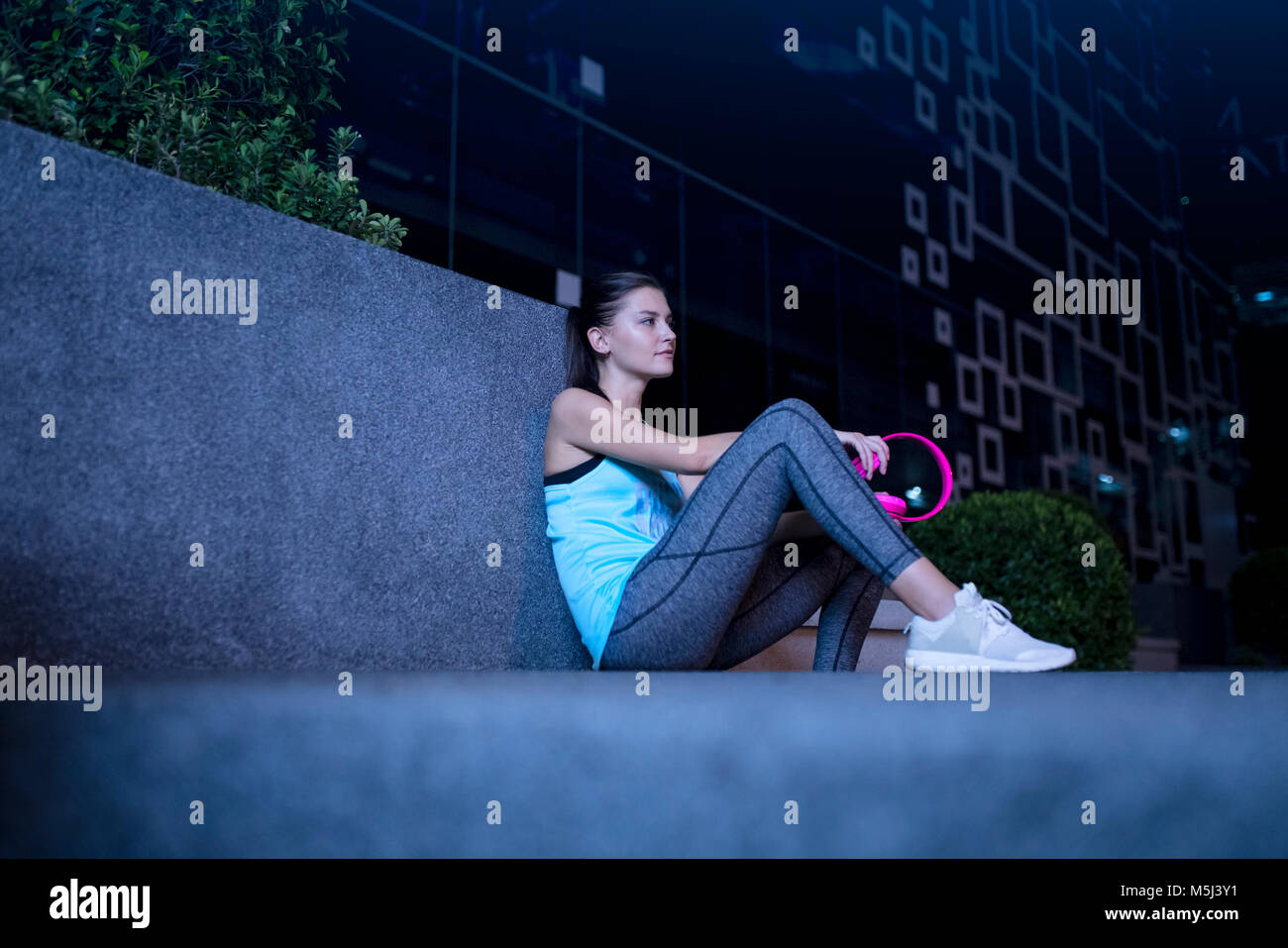 Young woman with pink headphones sitting on the ground in modern urban setting at night Stock Photo