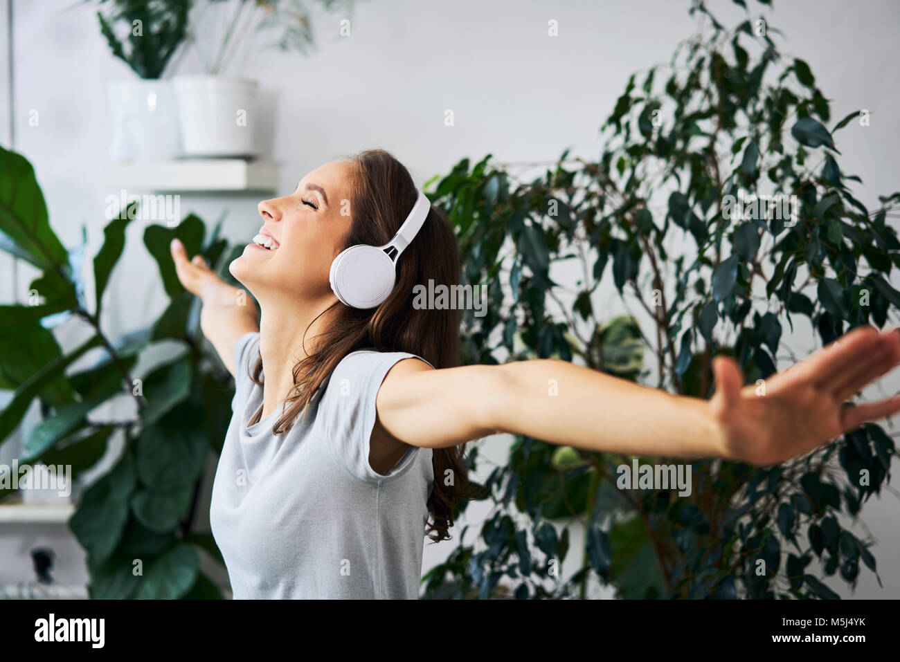 Happy young woman with oustretched arms listening to music at indoor plant Stock Photo