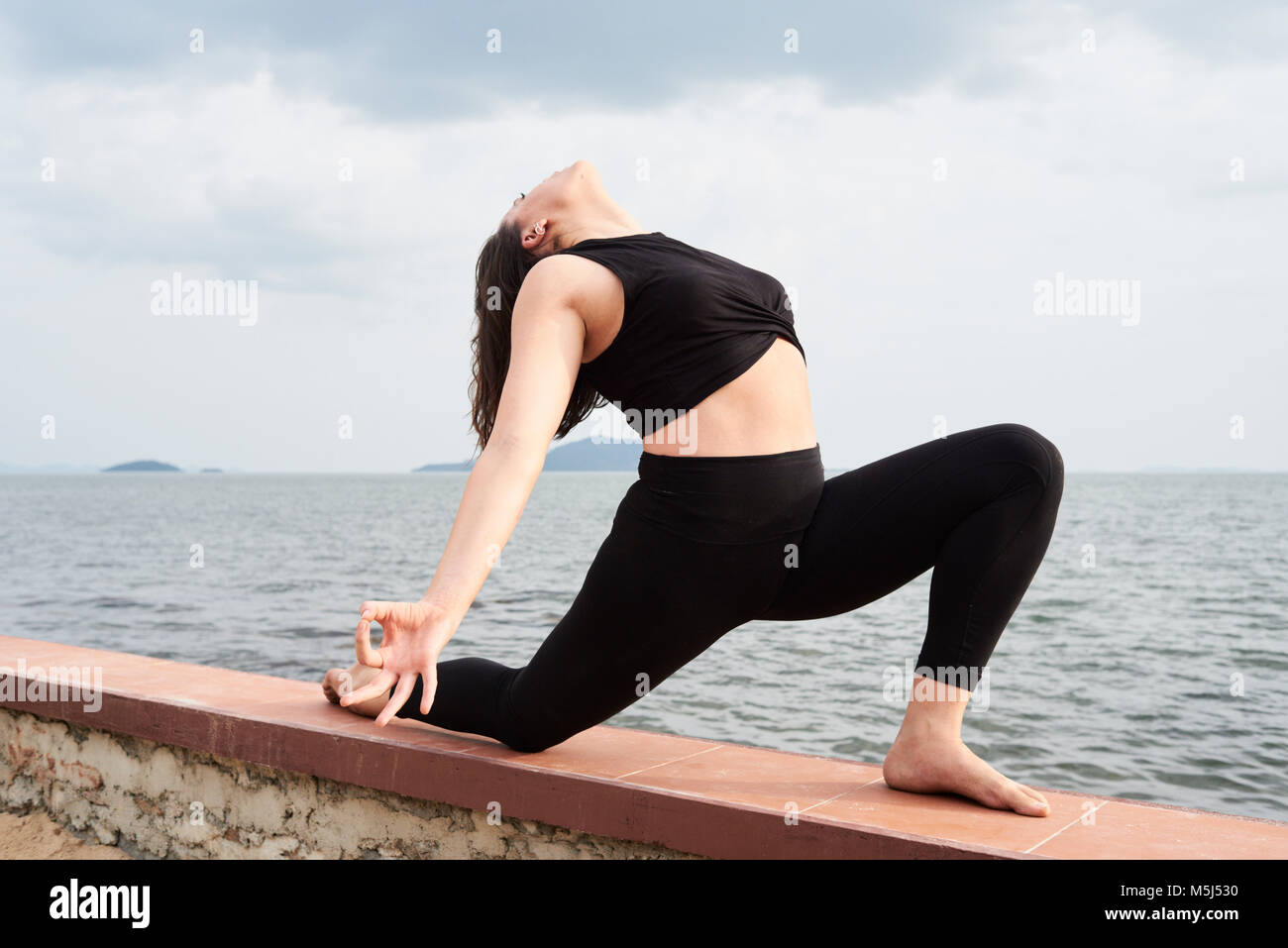 Yoga instructor in warrior position with arms opened and face up on a fence next to the sea. Kep, Cambodia. Stock Photo