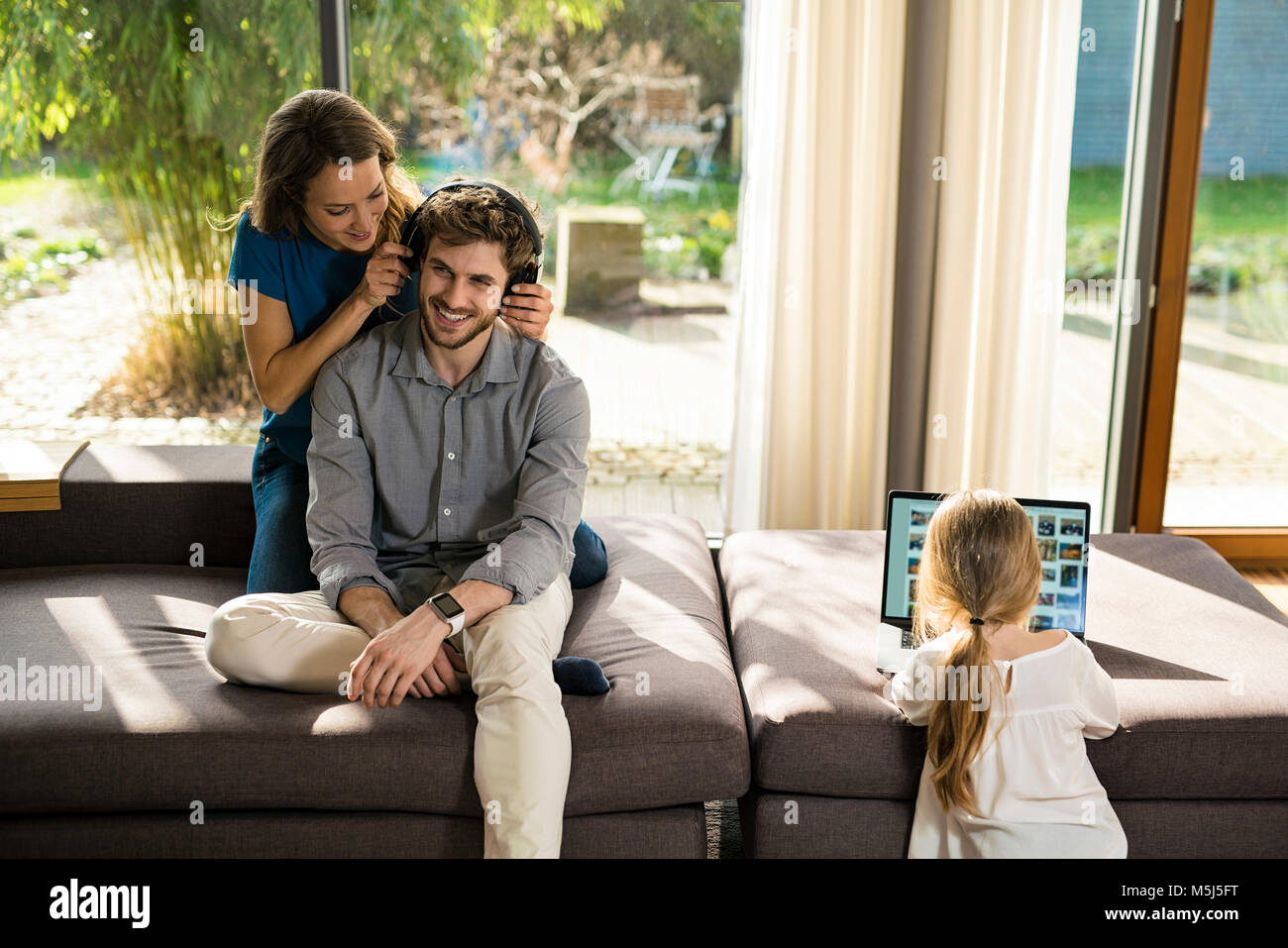 Couple with heaphones and daughter with laptop on sofa at home Stock Photo
