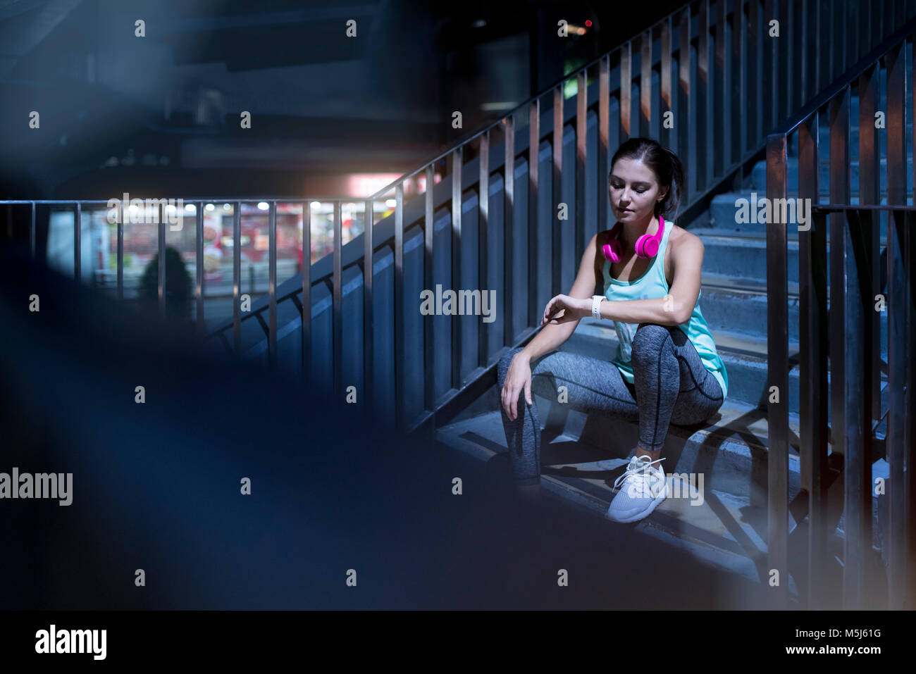 Young woman with pink headphones sitting on stairs and checking her smartwatch Stock Photo