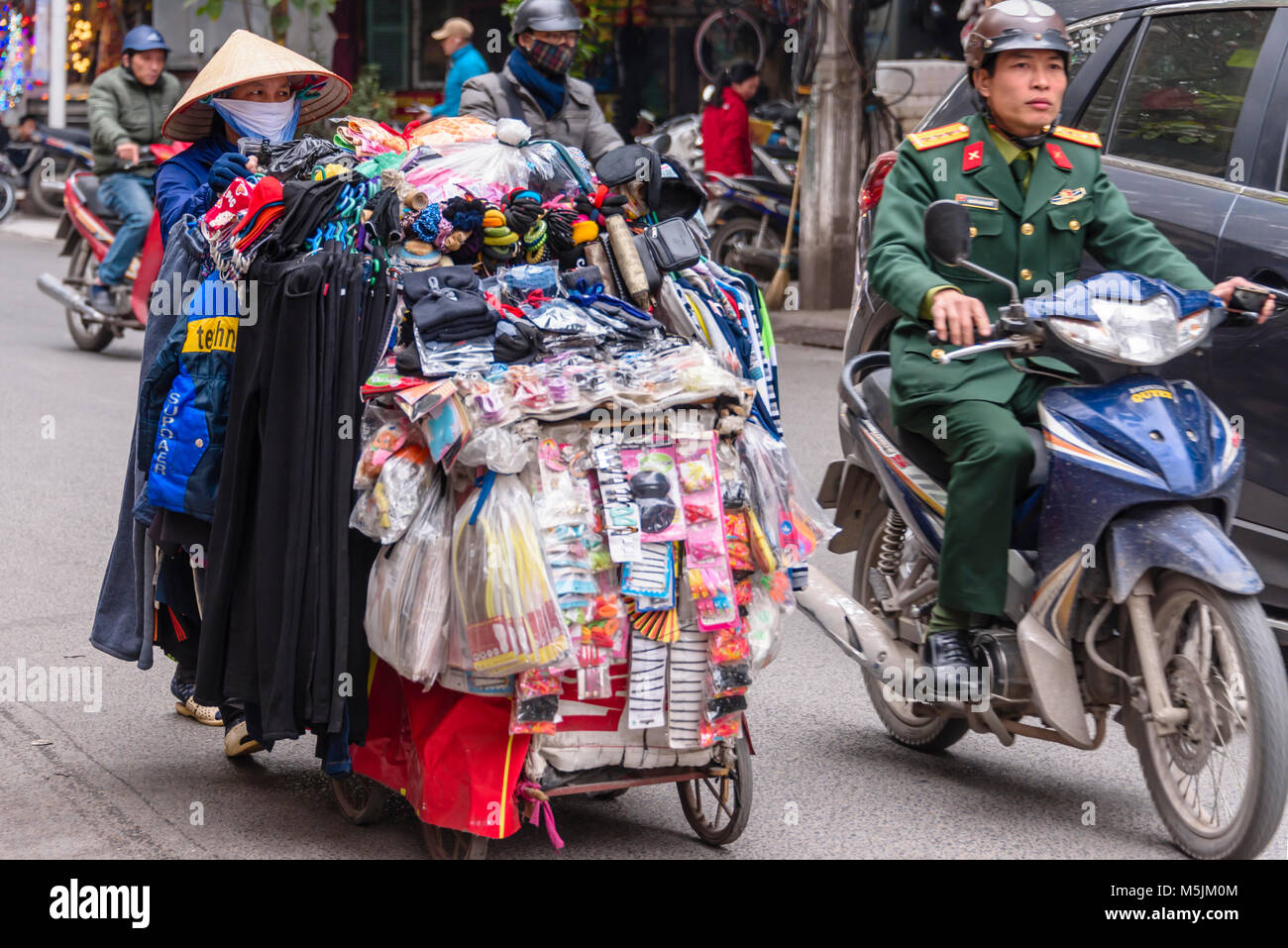 A Vietnamese woman wearing a bamboo hat and face mask, pushes a handcart full of household items for sale in Hanoi, Vietnam. Stock Photo