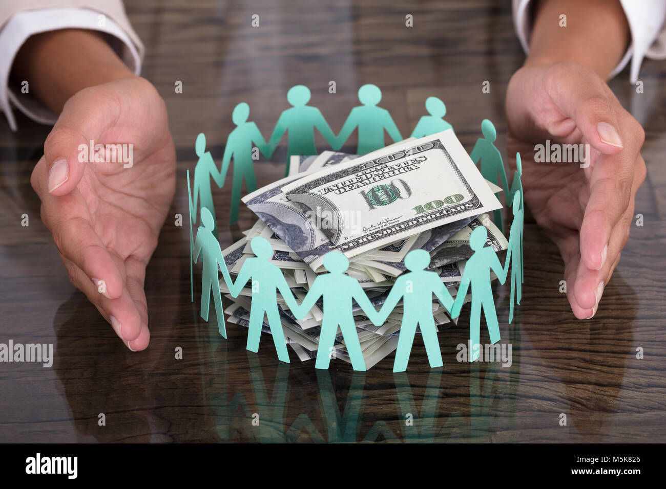Hand Protecting Paper Cut Out Human Figures Around Stack Of Hundred Dollar Bills Stock Photo