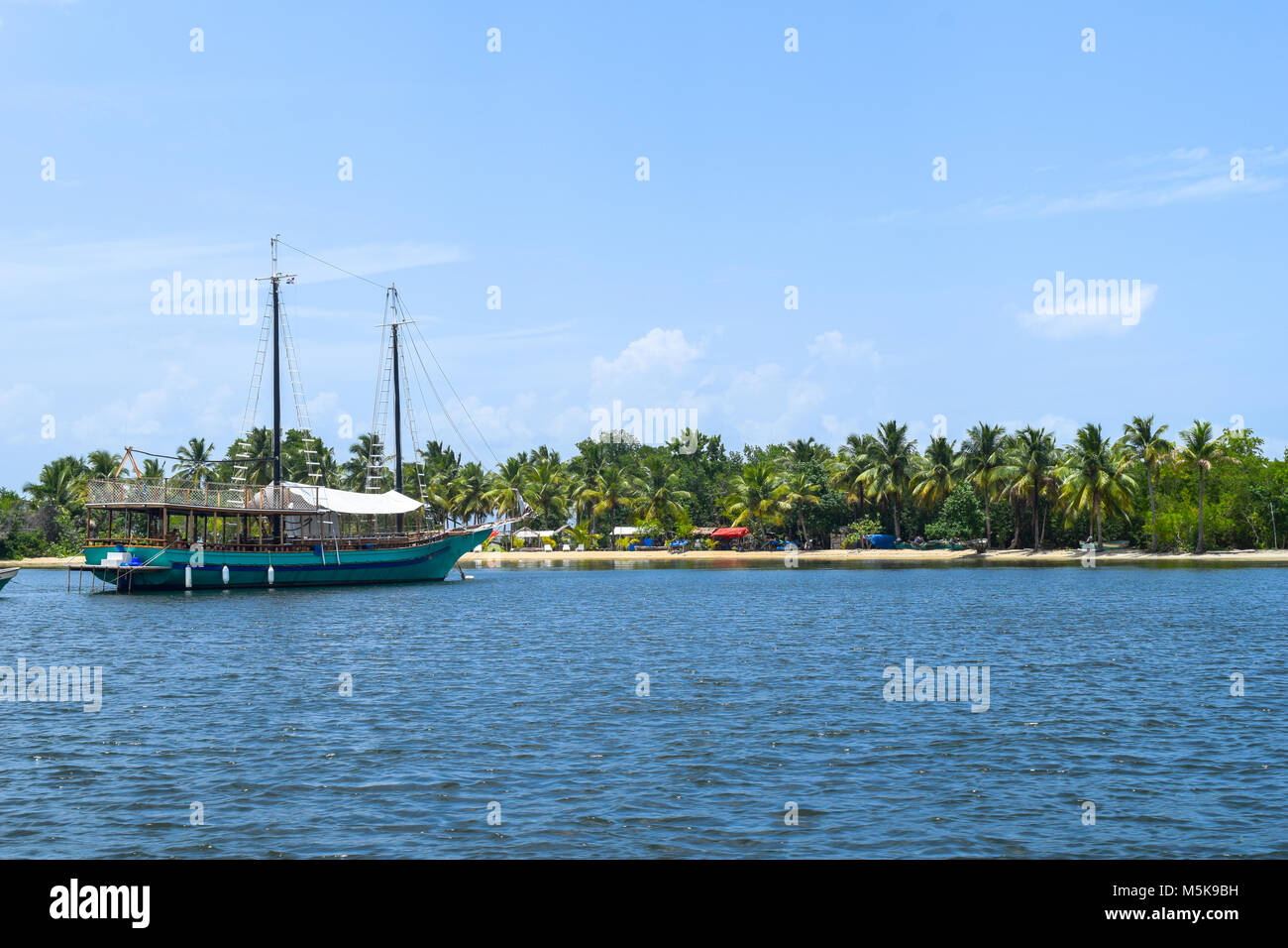 Excursion boat is waiting for the tourists in front of a beautiful tropical island with a white beach, sunbeds and many palms Stock Photo