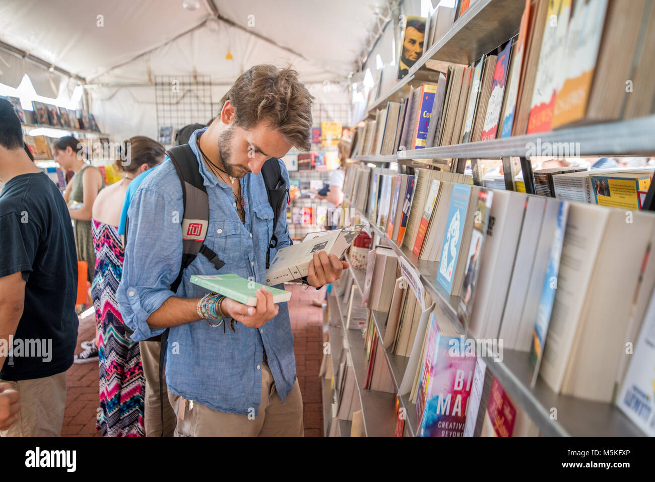 Young man reads the back of two books standing in front of bookshelf, Baltimore, Maryland. Stock Photo