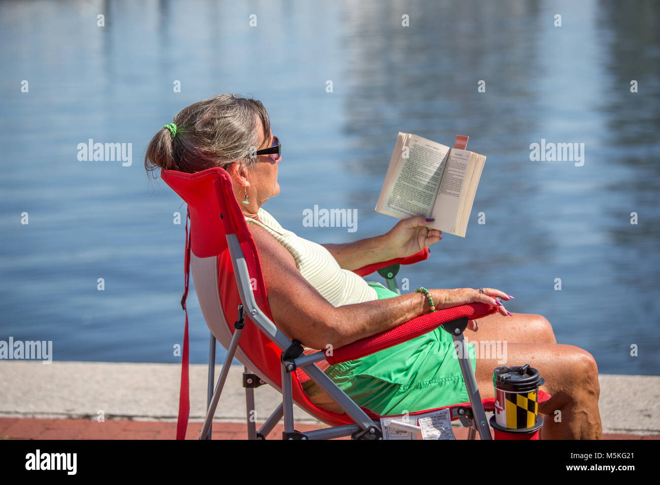 Middle aged woman relaxes in beach chair next to the water as she reads a book, Baltimore, Maryland. Stock Photo