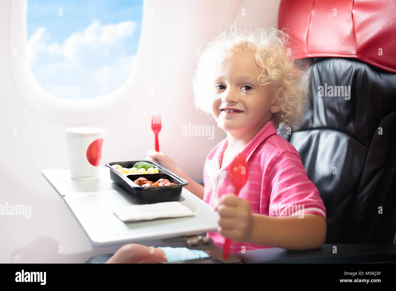 Child in airplane window seat. Kids flight meal. Children fly. Special inflight menu, food and drink for baby and kid. Little boy eating healthy lunch Stock Photo