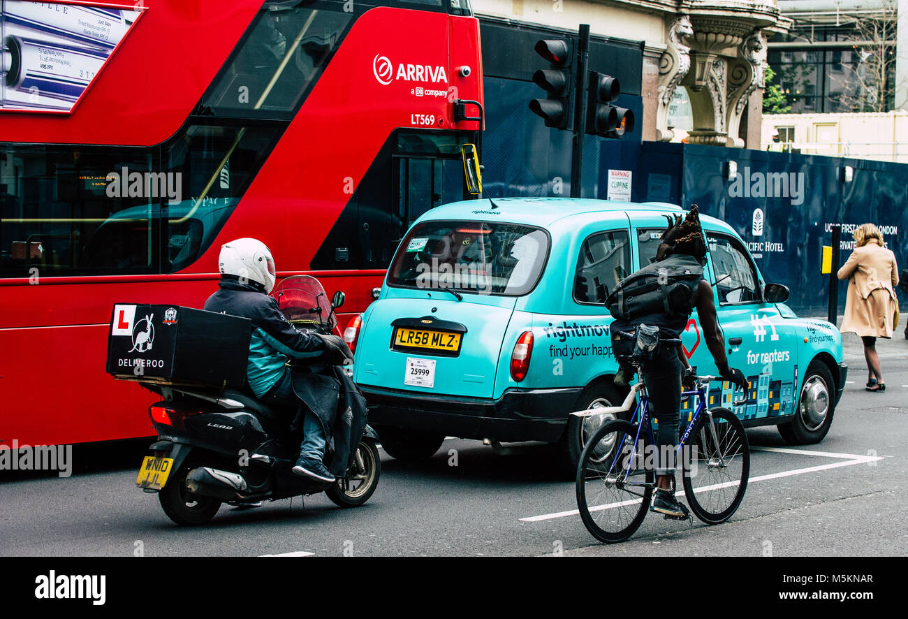 A deliveroo scooter, black cab taxi, cyclist and red London bus are lined up waiting at the traffic lights. Stock Photo