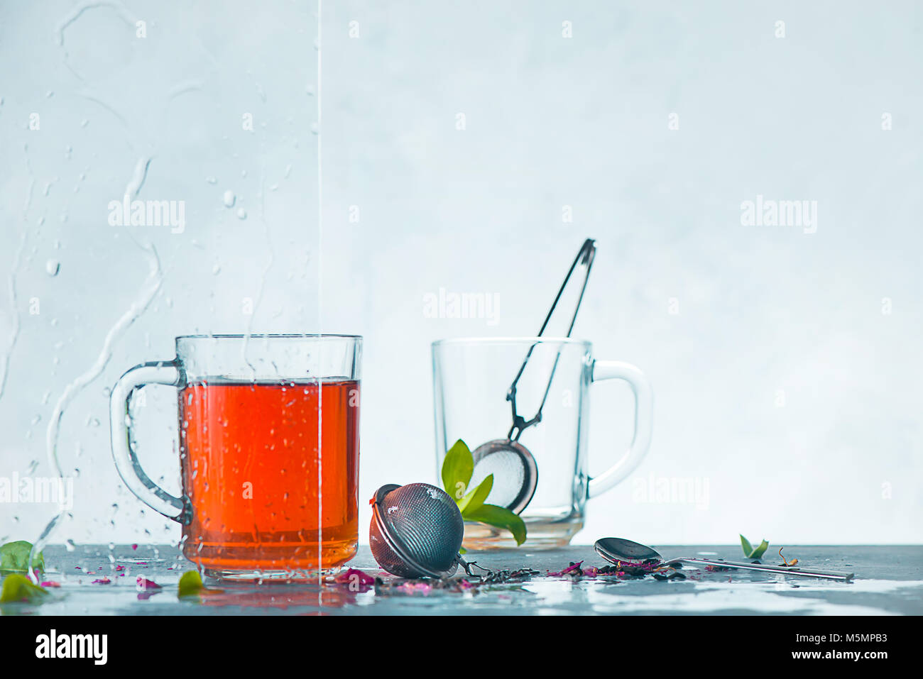 Cup of tea and cup with tea strainer, transparent on a light background with green leaves. Spring still life with raindrops on a window. Header with copy space. Stock Photo