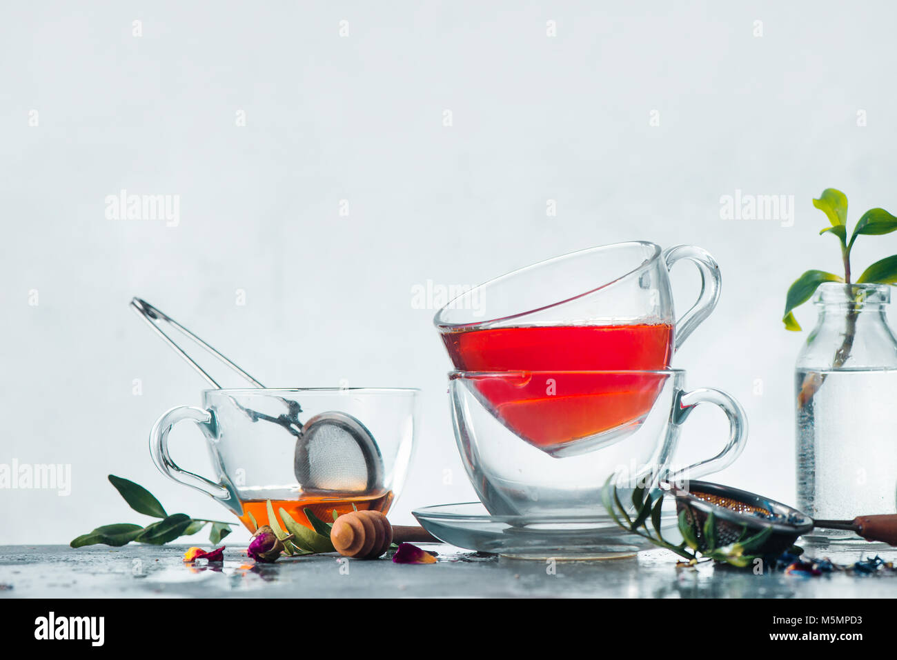 Stack of glass cups of tea on a white background with a honey spoon, tea strainer, fresh green leaves, flowers and copy space. Brewing herbal tea concept. High-key spring still life with copy space. Stock Photo