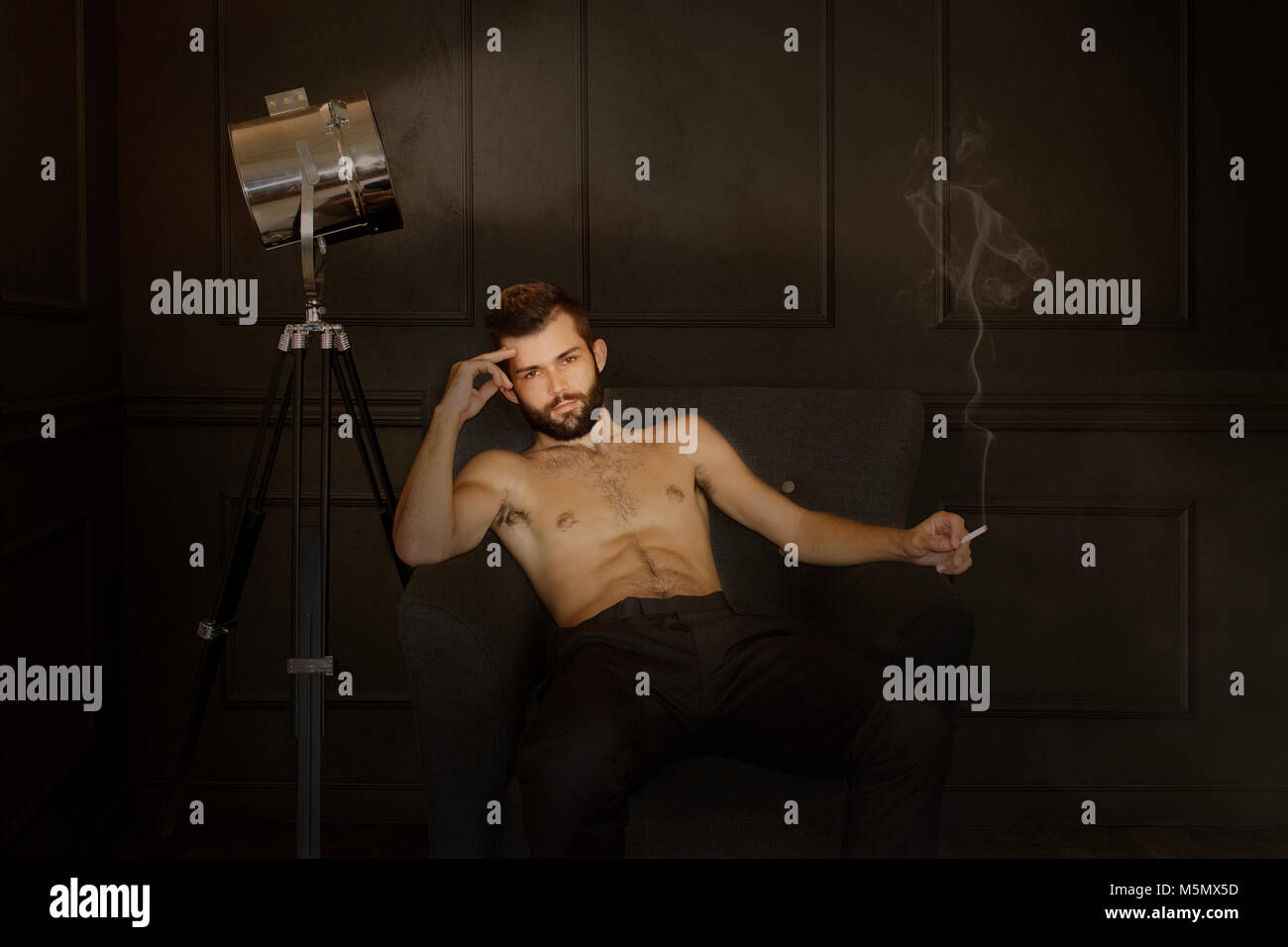 Young shirtless male actor reclines in a moody, smoke filled room while smoking a cigarette Stock Photo