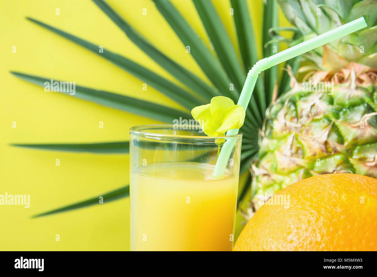 Close up of Tall Glass with Freshly Pressed Pineapple Orange Coconut Juice Straw and Small Flower. Round Palm Tree Leaf on Yellow Background. Vacation Stock Photo