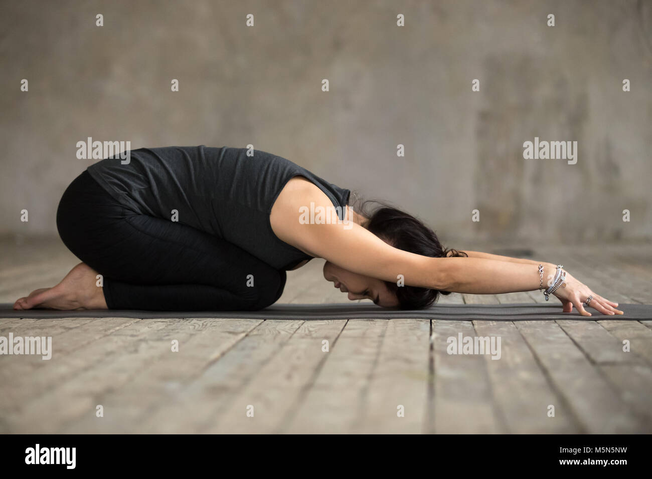 Young woman practicing yoga, doing Child exercise, Balasana pose, working out, wearing sportswear, black pants and top, indoor full length, gray wall  Stock Photo