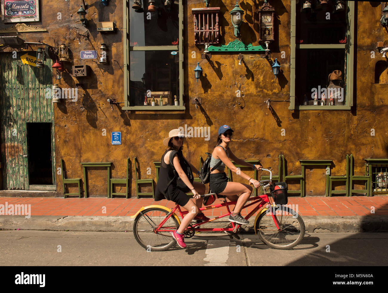 Two women on a tandem bicycle cycle past a colourful wall of a restauran in Cartagena, Colombia. Stock Photo
