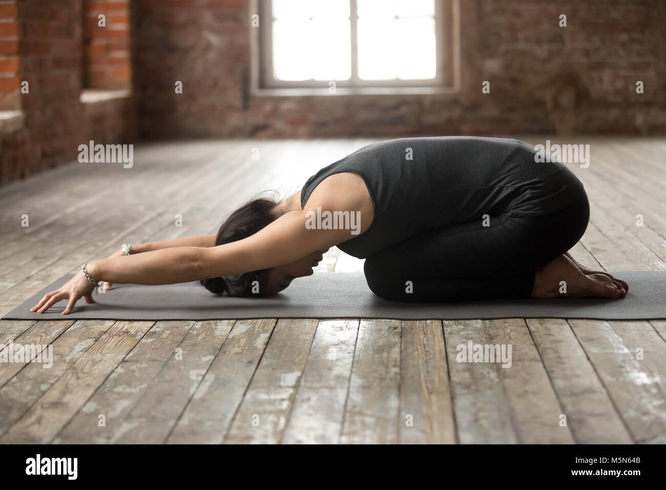 Young sporty woman practicing yoga, doing Balasana exercise, Child pose, working out, wearing sportswear, black pants and top, indoor full length, yog Stock Photo