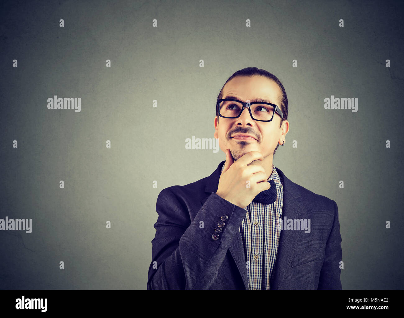 Young happy man in suit and glasses rubbing chin and daydreaming looking away on gray. Stock Photo