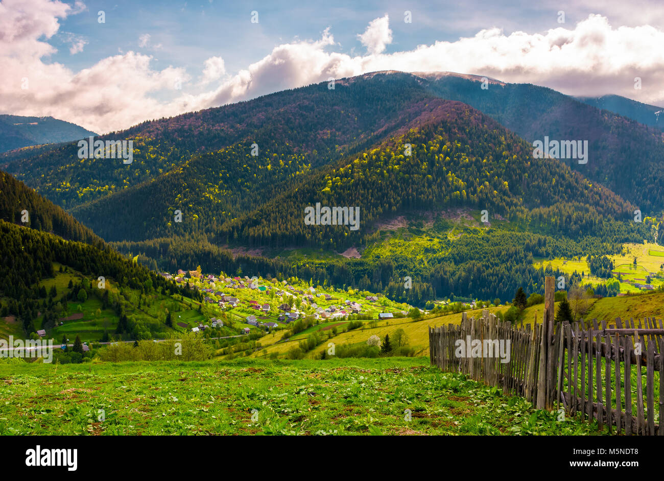 village in valley on a cloudy springtime day. fence on hillside above beautiful rural area Stock Photo