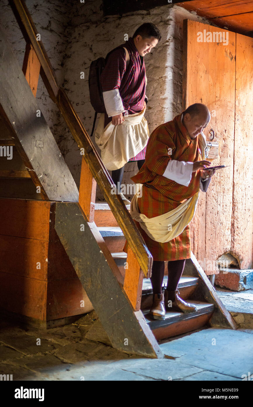 Jakar, Bhutan.  Men in Traditional Male Attire (Gho) Descending Stairway inside the Jakar Dzong (Monastery), One Using his Cell Phone. Stock Photo