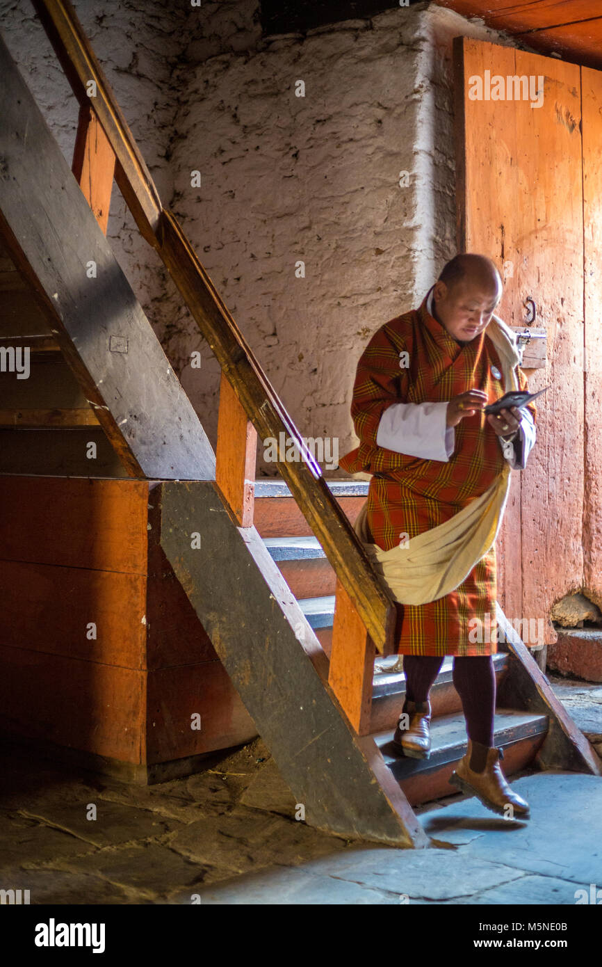 Jakar, Bhutan.  Man in Traditional Male Attire (Gho) Descending Stairway inside the Jakar Dzong (Monastery), Using his Cell Phone. Stock Photo
