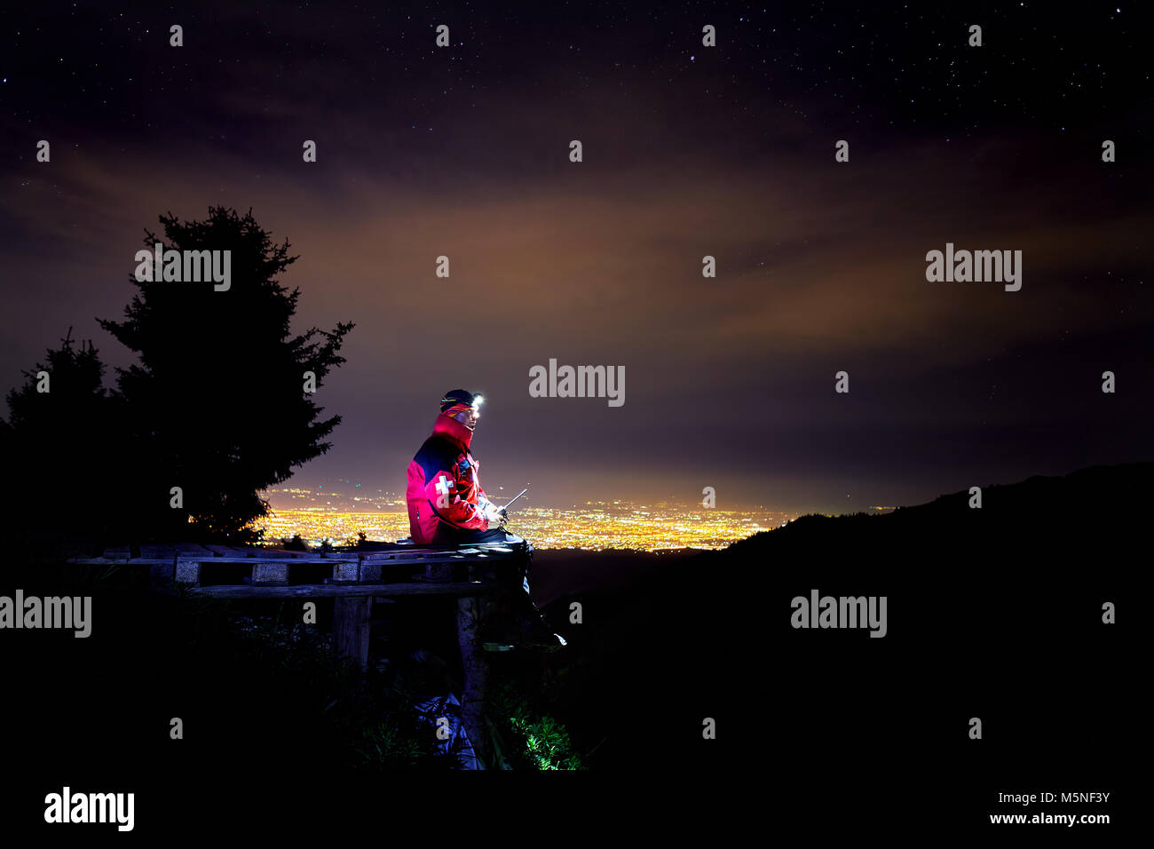 Lifeguard in red jacket with medical cross siting on the wooden branch in the mountain ski resort with view to night city light Stock Photo