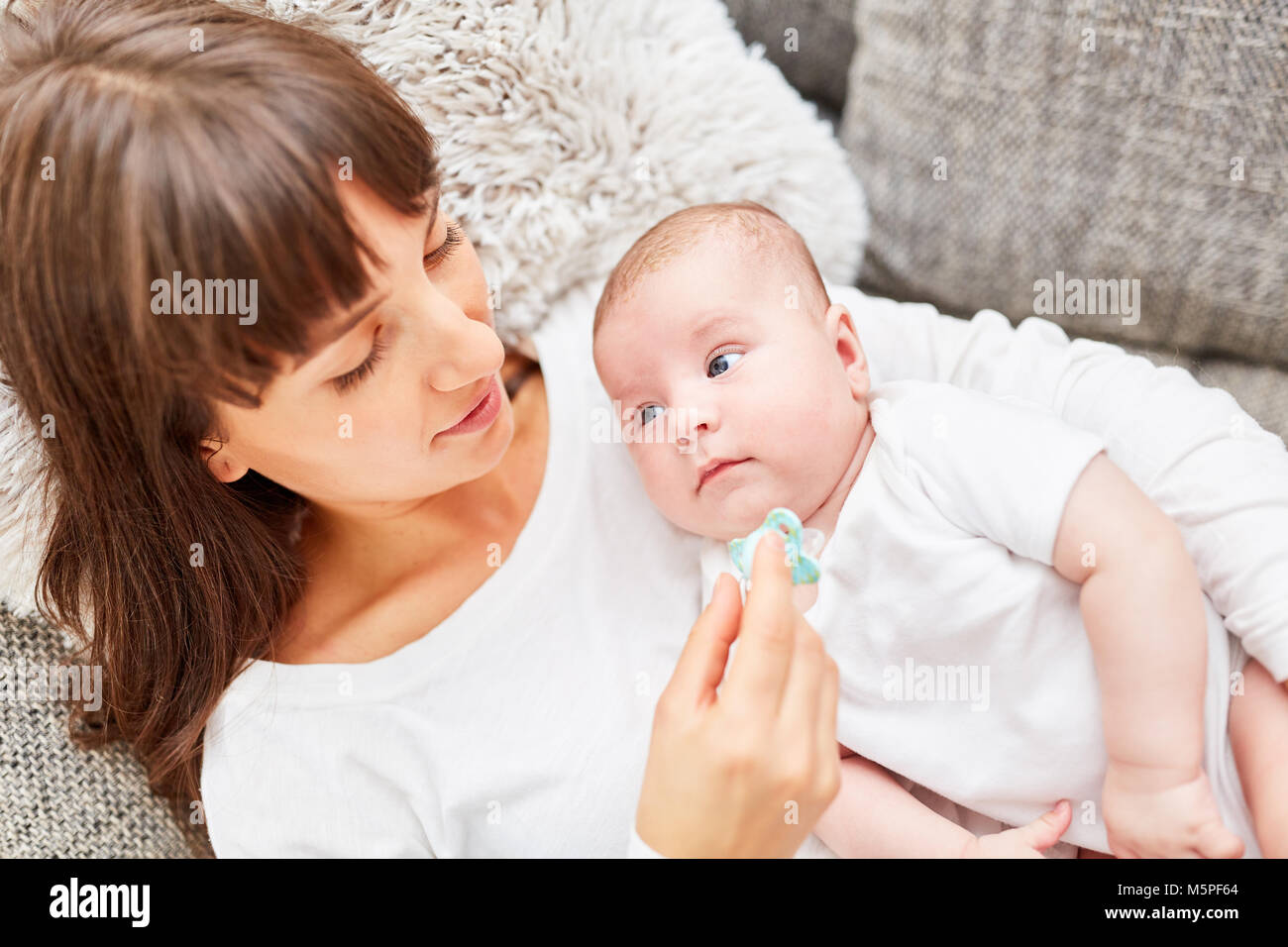 Happy mother with pacifier in hand together with baby on the sofa Stock Photo