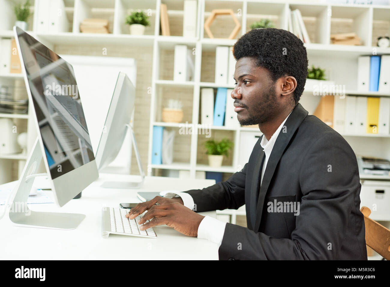 Profile view of confident African American manager sitting at desk and working on promising project with help of computer, interior of modern office o Stock Photo