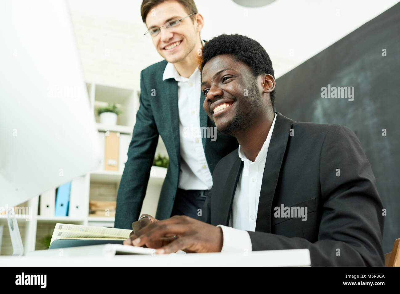 Multi-ethnic team of programmers with wide smiles gathered together in open plan office and working on start-up project with help of computer Stock Photo