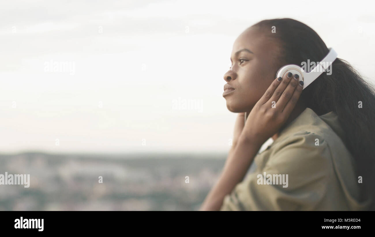 Side portrait of the beautiful attractive young african girl with natural make-up listening to music in headphones at the blurred background of the city. Stock Photo
