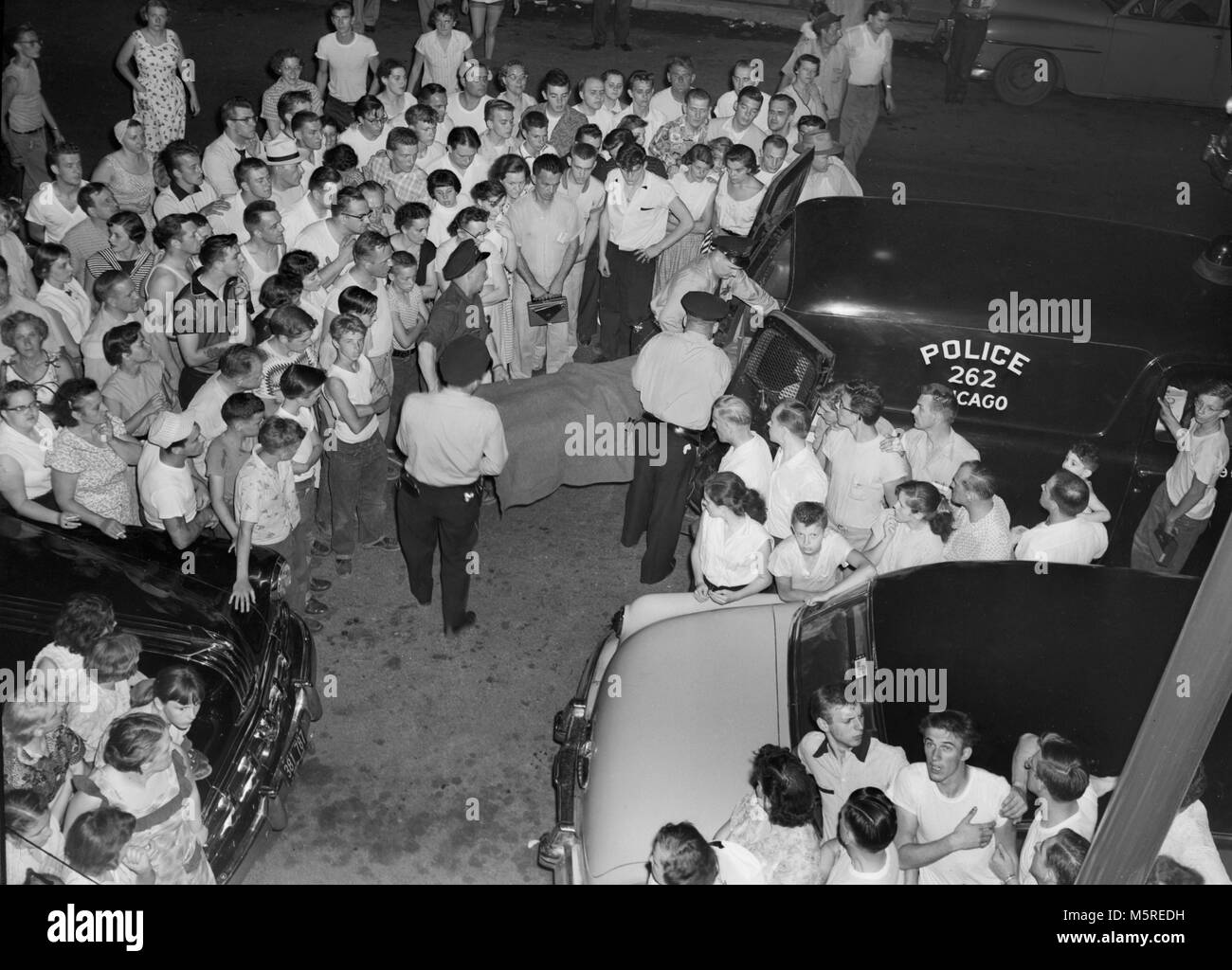 A neighborhood crowd watches as the body of murder victim is loaded in the back of police hearse in the South Side of Chicago, ca. 1955. Stock Photo