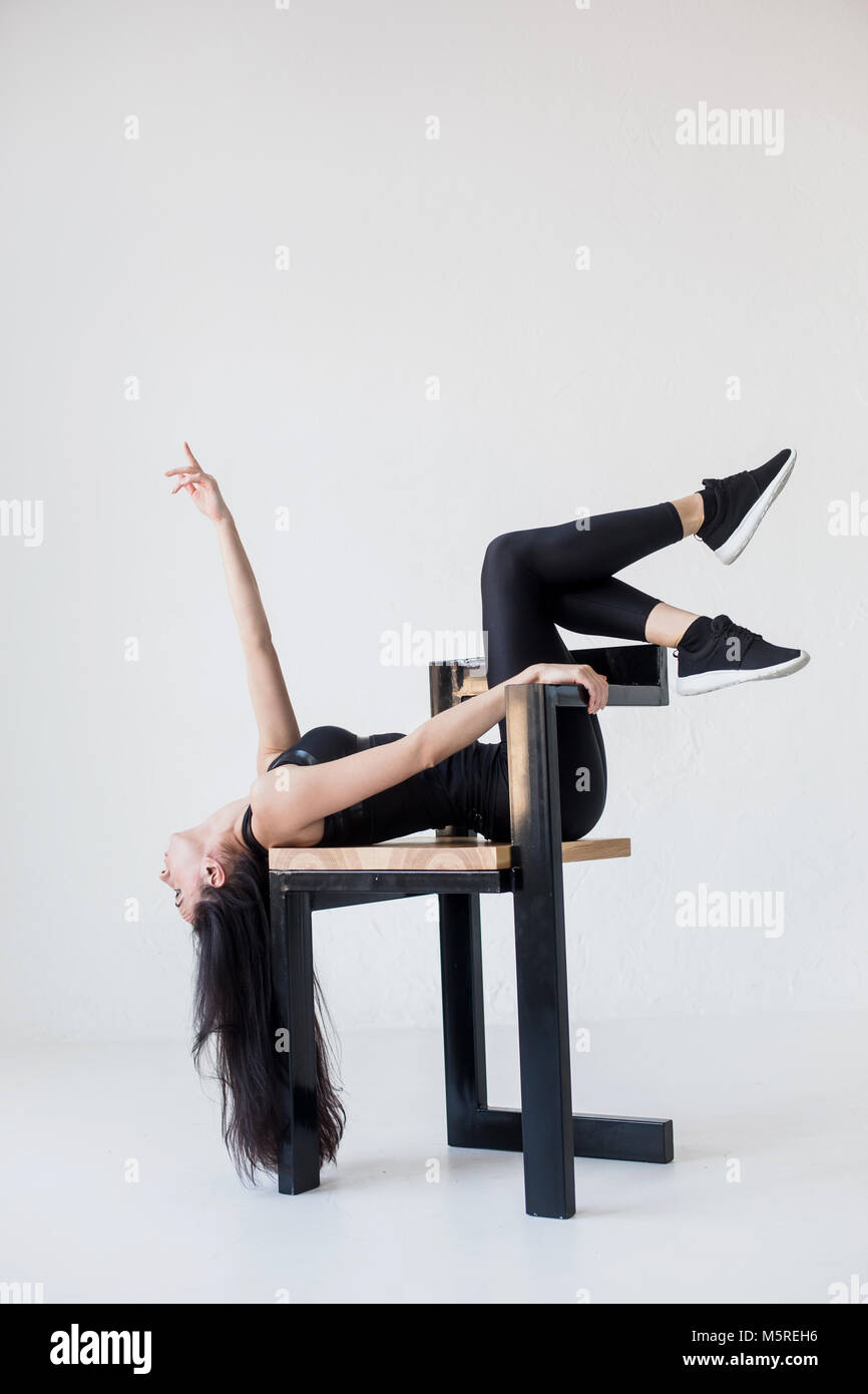 The sports attractive flexible woman in sportwear is stretching on the chair with the legs on the chair back at the white background. Stock Photo