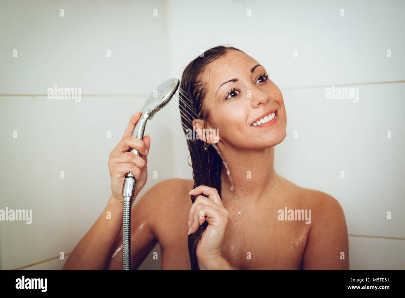 Happy young woman wetting her hair while showering under shower head. Stock Photo