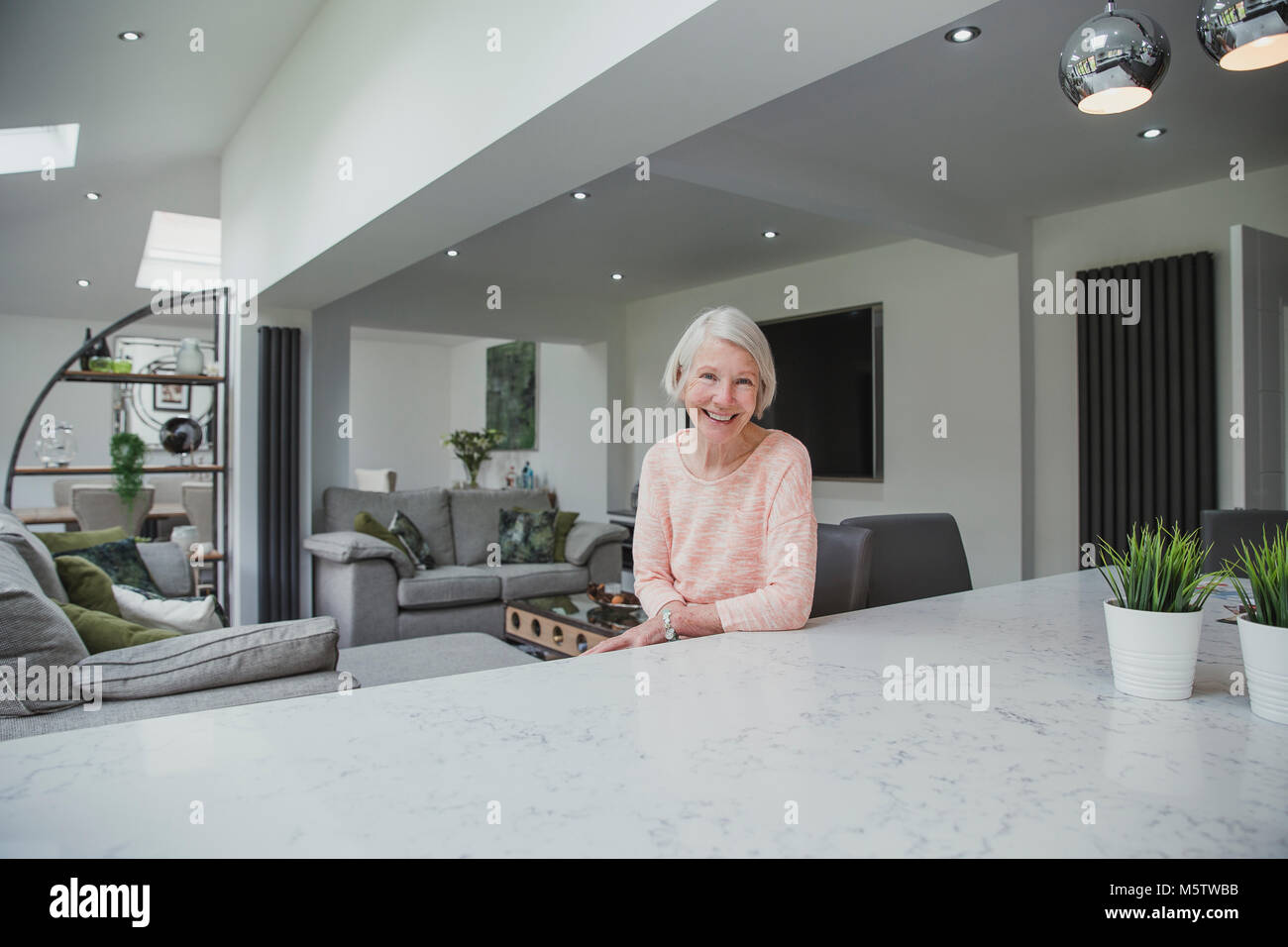 Happy senior woman is smiling for the camera while sitting at the breakfast bar in the kitchen of her home. Stock Photo
