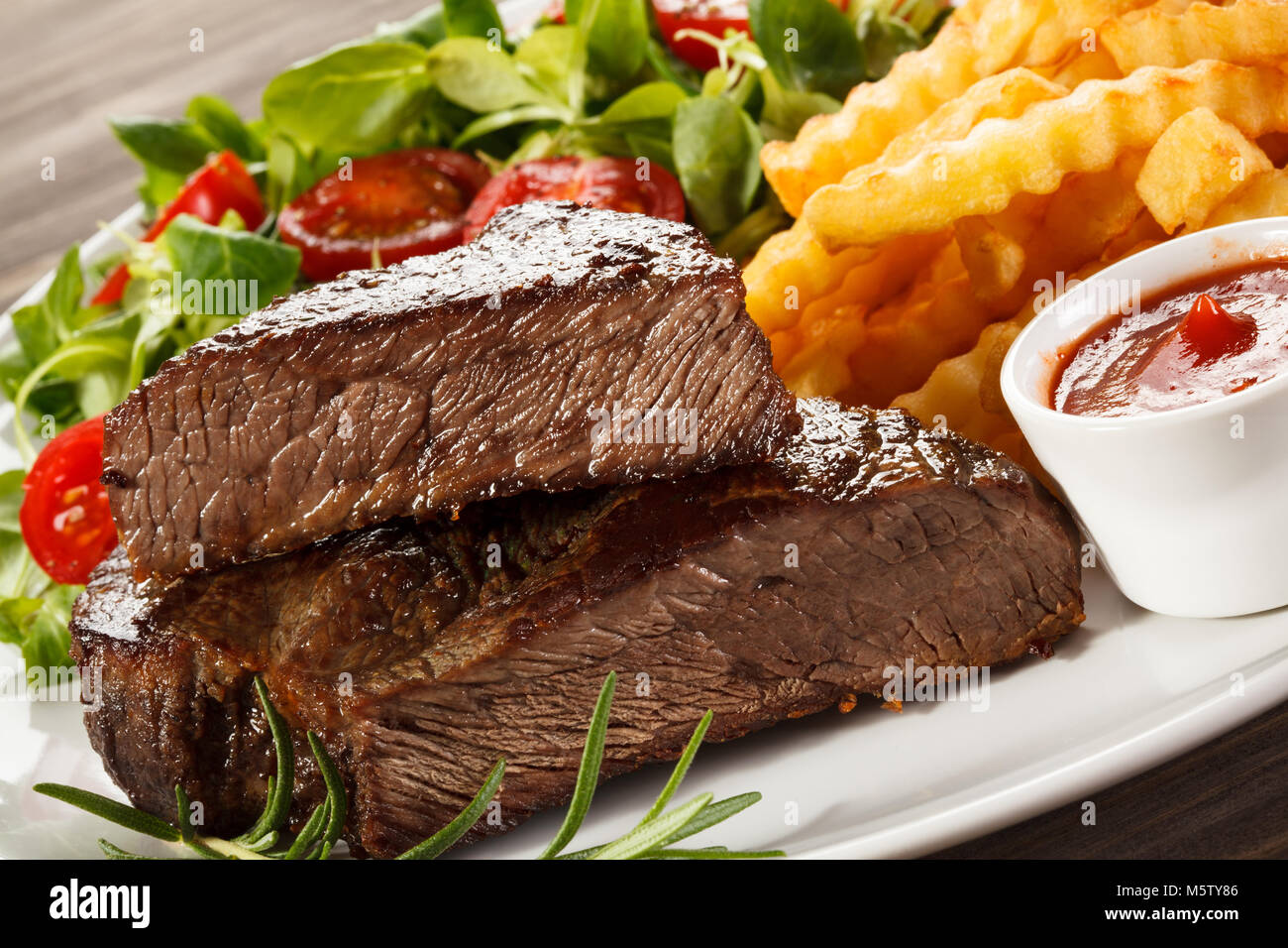 Grilled steak, French fries and vegetables on wooden background Stock Photo