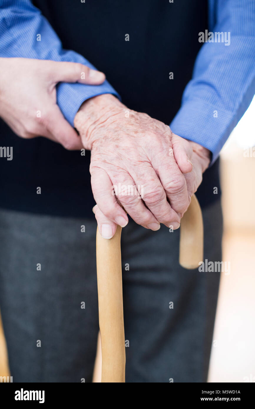 Senior Man's Hands On Walking Stick With Care Worker In Background Stock Photo