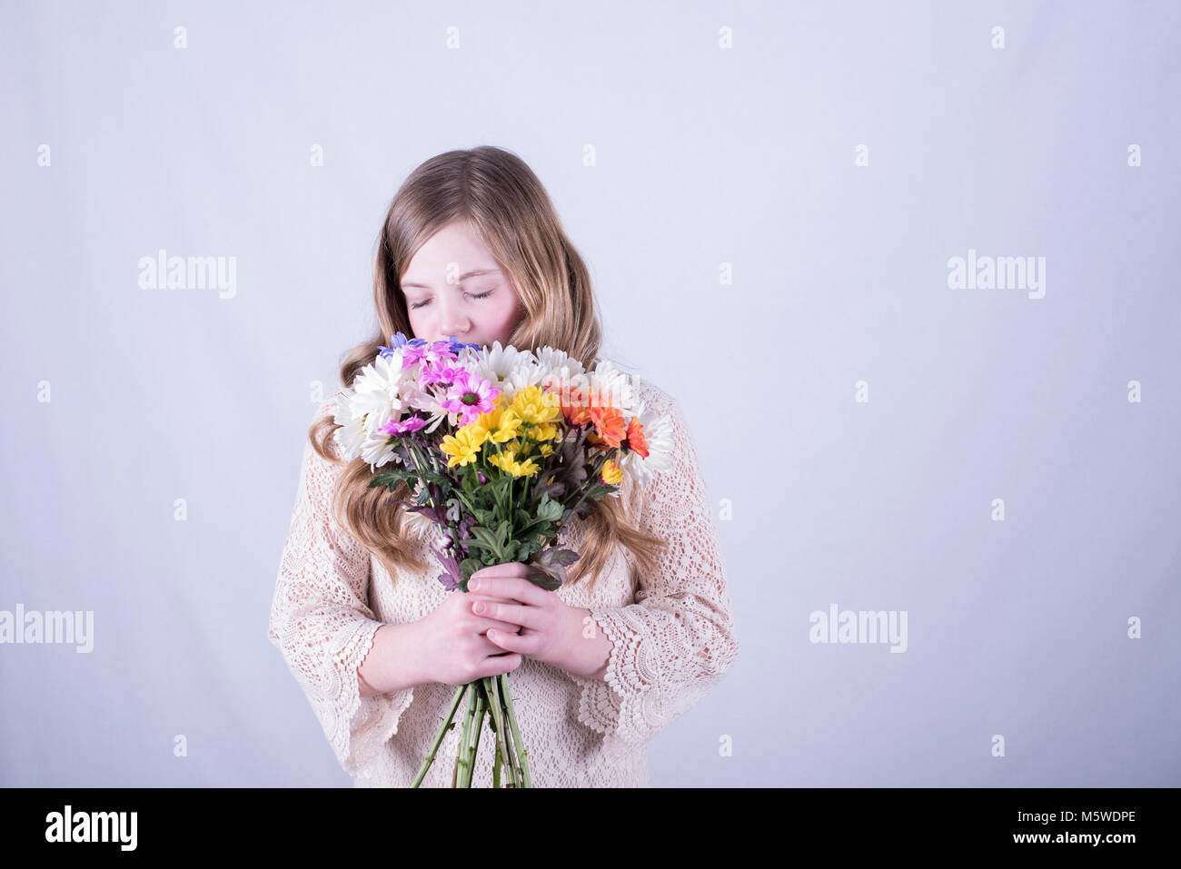 Twelve-year-old girl with long, dirty blonde hair holding colorful bouquet of daisies over lower face and smelling with eyes closed, white background Stock Photo