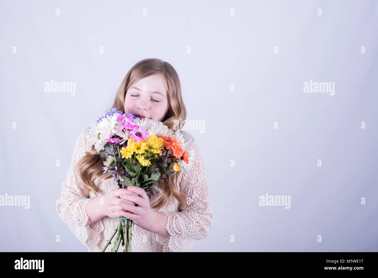Smiling twelve-year-old girl with long, dirty blonde hair holding and smelling colorful bouquet of daisies with eyes closed against white background Stock Photo