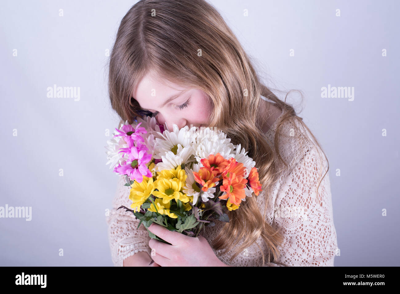 Smiling twelve-year-old girl with long, dirty blonde hair holding colorful bouquet of daisies and smelling with eyes closed against white background Stock Photo