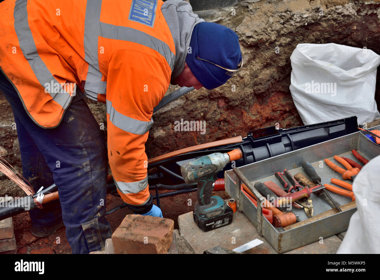 Electricity workman repairing underground mains electric cable working in hole in pavement Stock Photo